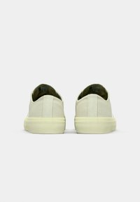 Scarpe slip-on in tela di colore beige chiaro, con una morbida texture, forma arrotondata e suole in gomma tono giallo. Dettagli di design minimalista.