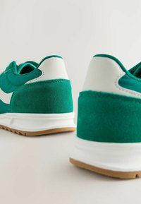 Baskets en suede vert avec des accents blancs, semelles rembourrées et détails texturés. Le design présente un bout arrondi et une semelle en caoutchouc.