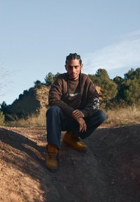 Smeđi dukserica s bijelim logom, crnim detaljima na rukavima, tamnoplave hlače i žute čizme. Prirodno okruženje s zemljanim tonovima.