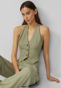 Oliwkowa bluzka na ramiączkach o kroju cropped, z zapięciem na guziki z przodu. Tkanina ma gładką fakturę i minimalistyczny styl.