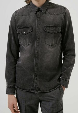 Muž v vybledlé černé džínové košili s knoflíkovými kapsami na hrudi a tmavých károvaných kalhotách stojí s jednou rukou v kapse.