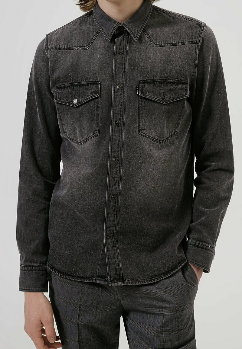 Homme portant une chemise en jean noire délavée avec des poches poitrine boutonnées et un pantalon à carreaux foncés, debout avec une main dans la poche.