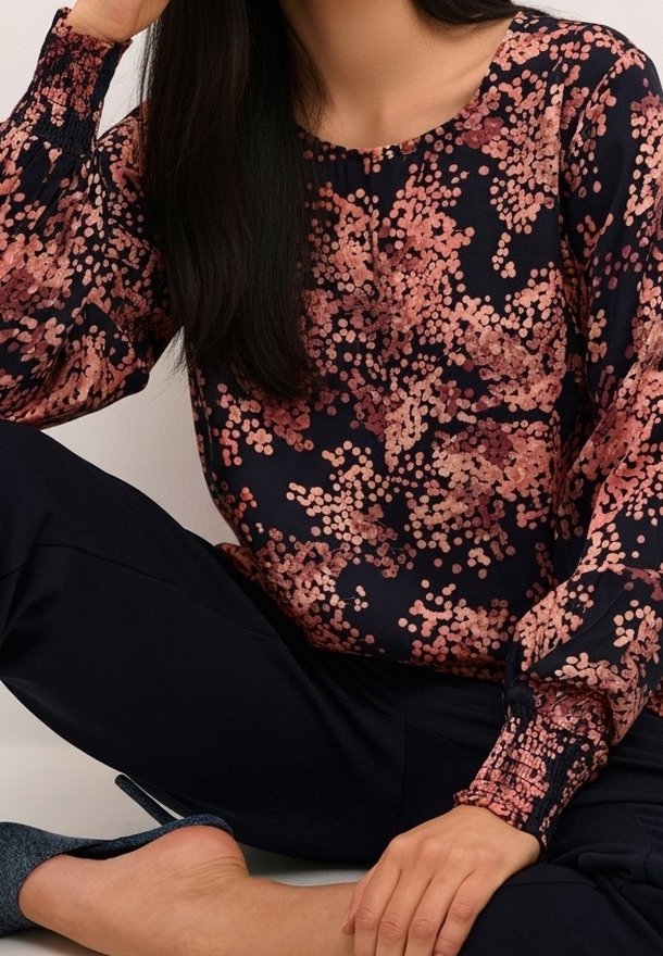 Femme assise en tailleur, portant un chemisier bleu marine à manches longues avec des pois floraux roses et un pantalon noir, ainsi que des ballerines bleues.