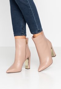 Nude skórzane botki za kostkę z szpiczastymi noskami i metalowymi obcasami w formie bloku. Posiadają boczny zamek błyskawiczny z zawieszką w kształcie serca. Gładka tekstura.