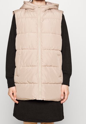 Veste sans manches matelassée beige à capuche avec fermeture éclair à l'avant portée sur une robe noire à manches longues côtelée.