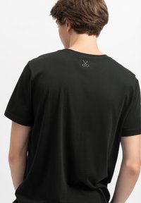 Zwarte T-shirt met korte mouwen van katoen, met een klein schaarlogo in een contrasterende kleur op de bovenrug. Gladde textuur.