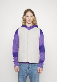 Gequiltete hellgraue Weste mit V-Ausschnitt, vorderem Knopfverschluss und glatter Oberfläche, getragen über einem lila Batik-Hoodie. Jeans runden das Outfit ab.
