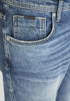 Vaqueros de mezclilla en azul claro con una textura desgastada. Cuentan con un bolsillo para monedas, costuras amarillas en contraste y una etiqueta negra en el bolsillo trasero.