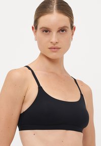 Woman with light skin and straight hair pulled back wearing a simple black sleeveless bralette, facing forward against a plain background.
