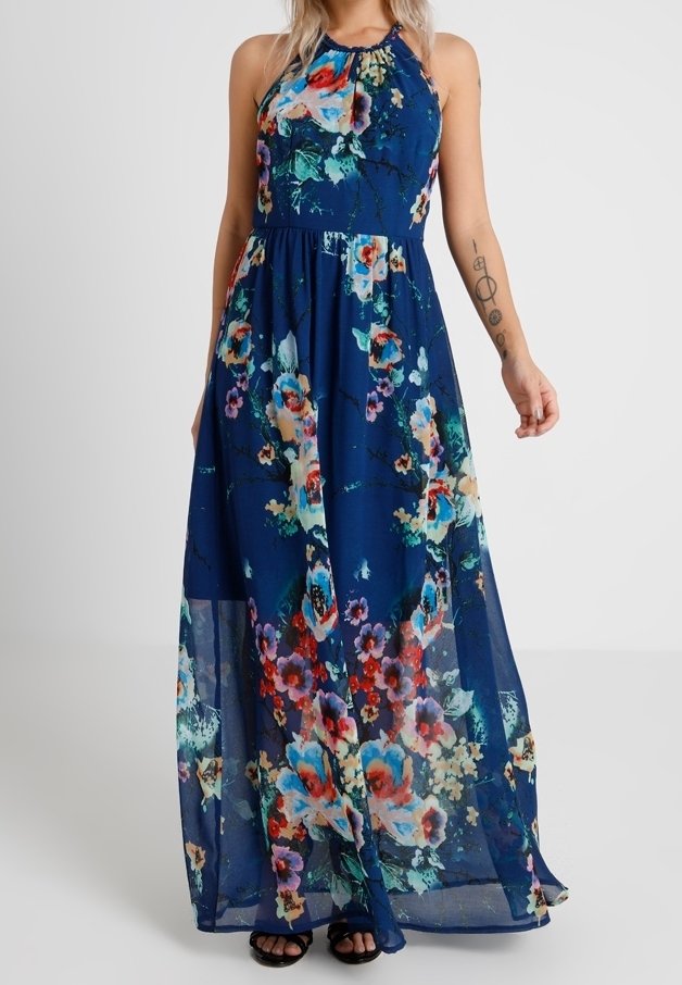 Femme portant une robe maxi bleu marine sans manches avec des motifs floraux colorés et des sandales noires, debout devant un fond uni.