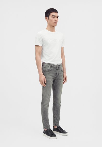 Jeune homme debout portant un t-shirt blanc, un jean gris ajusté et des baskets noires sur un fond clair uni.