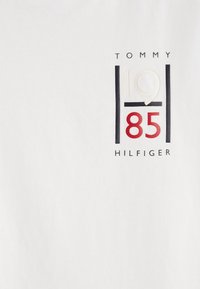 Camiseta blanca con un gráfico central que presenta "TOMMY", "1985" y "HILFIGER" en texto negro y rojo con acentos contorneados.