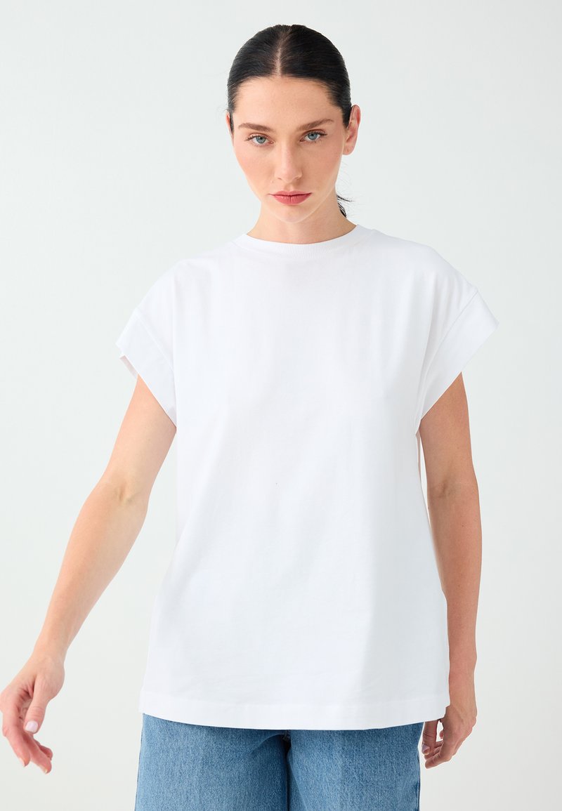 Femme aux cheveux foncés attachés porte un t-shirt blanc uni à manches courtes et un jean bleu, debout devant un fond clair.