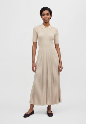 Une femme porte une robe en maille beige à manches courtes, avec un haut à col et une jupe midi plissée, assortie à des chaussures plates foncées.
