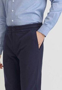 Donkerblauwe broek met een elastische tailleband, voorzien van een knoopsluiting, zijzakken en een soepele katoenen textuur. Draag met een lichtblauw shirt.