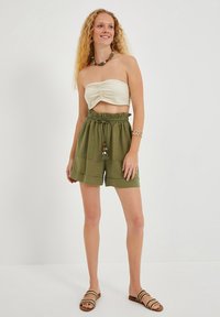 Top bandeau z kremowego materiału; oliwkowe szorty z elastycznym pasem i kieszeniami; brązowe sandały z paskami; naszyjnik z koralików.