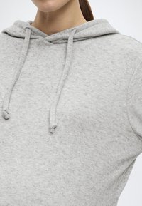 Gaiši pelēks džemperis ar kapuci no mīksta, teksturēta auduma. Aprīkots ar kengūras kabatu, kapuci ar aukliņu un brīvu piegriezumu.