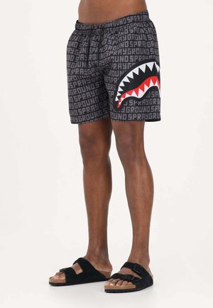 Uomo che indossa pantaloncini neri con scritta grigia "SPRAYGROUND" su tutta la superficie e un grande design della bocca di uno squalo rosso, bianco e nero sulla gamba destra.