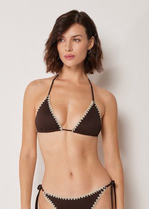 Frau mit kurzen, welligen Haaren, die einen dunkelbraunen, strukturierten Bikini mit cremefarbenem, geschwungenem Saum trägt, steht vor einem einfachen hellen Hintergrund.