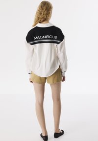 Camicia bianca con accenti neri, con la scritta "MAGNIFIQUE" sulla schiena; mostrata con pantaloni corti beige e scarpe nere slip-on.
