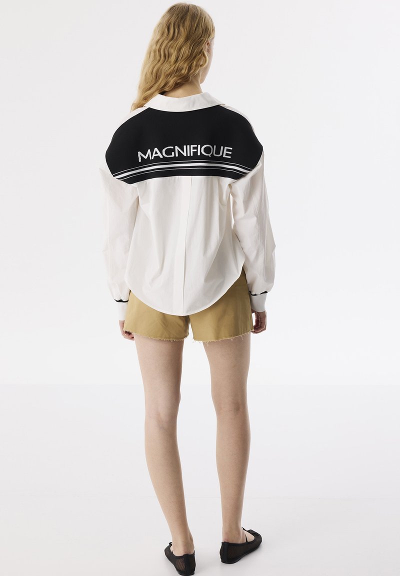 Camicia bianca con accenti neri, con la scritta "MAGNIFIQUE" sulla schiena; mostrata con pantaloni corti beige e scarpe nere slip-on.