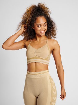 Femme aux cheveux bouclés portant un soutien-gorge de sport beige et des leggings, souriante et touchant ses cheveux sur un fond uni.