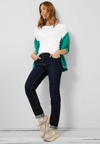 Witte langlopende overhemd, groene jas over de schouders gedrapeerd, donkere denim jeans en beige sneakers met een gestructureerd patroon.