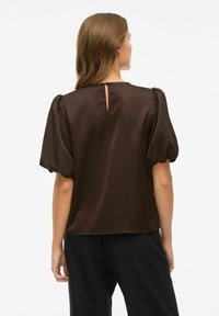 Vrouw met lichtbruin haar, gekleed in een donkerbruine blouse met pofmouwen en zwarte broek, staande met haar rug naar de camera tegen een effen witte achtergrond.