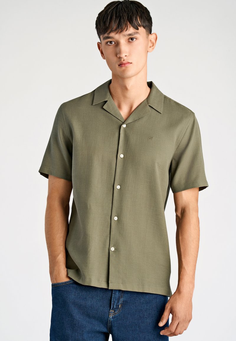 Młody mężczyzna w oliwkowej zielonej koszuli z krótkim rękawem i małym haftowanym logo, w połączeniu z niebieskimi dżinsami, pozujący na białym tle.