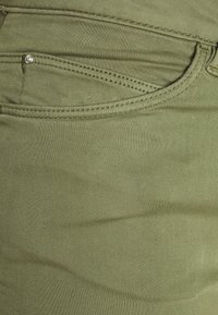 Groen katoenen stof met een gladde textuur, met een zak voorzien van een zilveren knoop en zichtbare stikseldetails.