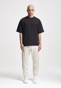 Schwarzes Oversized-T-Shirt mit kurzen Ärmeln, kombiniert mit hellbeigen Jogginghosen und weißen Sneakers, vor einem schlichten Hintergrund.