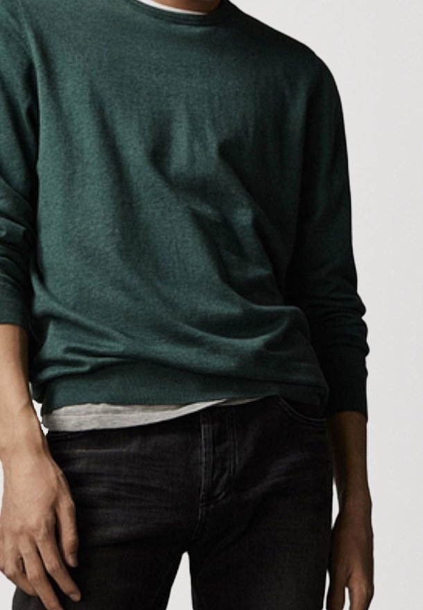 Homme portant un pull vert foncé superposé à une chemise gris clair et un jean noir, sur un fond gris uni.