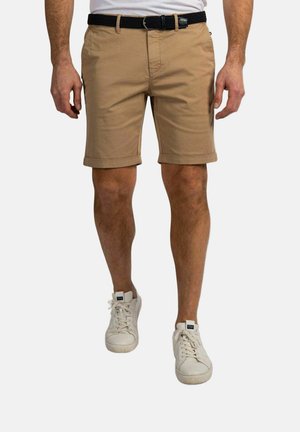 Man draagt beige knielange shorts met een zwarte riem en witte sneakers, staand tegen een effen achtergrond.