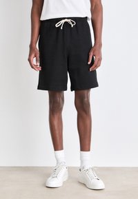 Black sweat shorts with an elastic waistband and beige drawstring, featuring a relaxed fit and rolled hem. Worn with white sneakers.