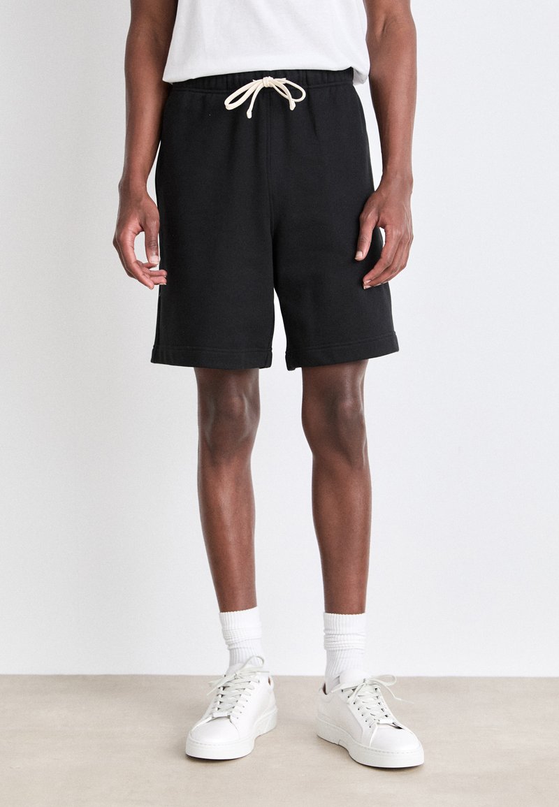 Black sweat shorts with an elastic waistband and beige drawstring, featuring a relaxed fit and rolled hem. Worn with white sneakers.