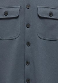 Pelēka teksturēta krekla ar pogām, kas piedāvā divas krūšu kabatas un melnas pogas. Audumam ir smalka herringbone raksts.