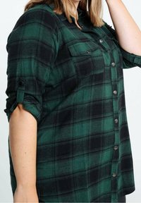 Zielono-czarna flanelowa koszula w kratę z długimi rękawami, zapinana na guziki z przodu, z dwoma kieszeniami na piersi, rękawy podwinięte i przypięte na guziki.