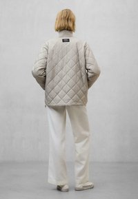 Hellbeige, gesteppte Jacke mit hohem Kragen und schwarzem Etikettendetail, kombiniert mit weiten weißen Hosen und hellen Schuhen. Rückansicht.