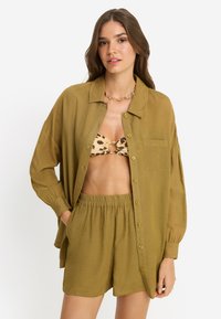 Πουκάμισο χρώματος ελιάς με κουμπιά, χαλαρή εφαρμογή και μακριά μανίκια, συνδυασμένο με σορτς στο ίδιο χρώμα· διαθέτει τσέπη και απαλή υφή.