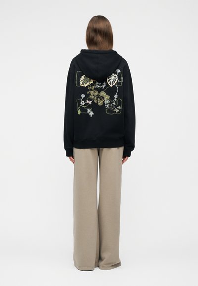 Άτομο που φοράει μαύρο φούτερ με μοτίβο φύλλων σε εκρού και πράσινο χρώμα στην πλάτη, συνδυασμένο με φαρδιά μπεζ παντελόνια, στέκεται μπροστά σε ένα απλό φόντο.