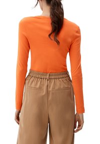 Maglietta arancione a maniche lunghe abbinata a pantaloni beige con vita elasticizzata, dotati di due tasche posteriori, realizzati in tessuto morbido e leggero.