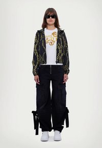 Črna jakna z vzorcem zlatih verig, bela majica s potiskom z logotipom, črne cargo hlače z žepi, zavezani detajli in beli supergi.