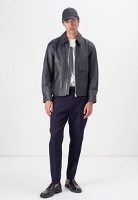 Jachetă din piele neagră cu fermoar în față, două buzunare frontale, combinată cu pantaloni eleganți bleumarin și pantofi negri, sub o șapcă întunecată.
