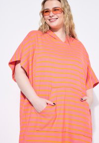 Tunic vestido con capucha en tonos rosa y naranja, con rayas, mangas cortas y un bolsillo frontal. Textura suave, corte holgado y diseño casual.