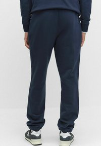 Person i mörkblå joggers och grå New Balance-sneakers, stående mot en enkel ljus bakgrund, sedd bakifrån.