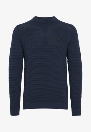 Tummansininen teksturoitu neule, jossa on pyöreä kaula-aukko ja ribbireunat. Tyylissä on pieni musta logo-etiketti rinnassa.