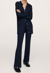 Osoba v tmavě modrém žebrovaném dlouhém cardiganovém svetru s knoflíky a odpovídajících rozšířených kalhotách, s černými mokasíny, stojící se zkříženýma rukama.