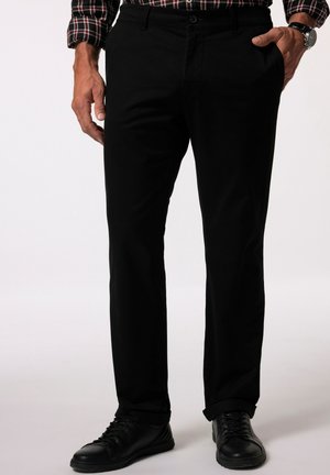 Homme portant un pantalon noir, des baskets en cuir noir, une chemise à manches longues à carreaux, main gauche dans la poche, main droite détendue le long du corps.