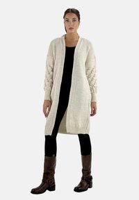 Lång, öppen cardigan i krämfärg, med texturerade ärmar och ett diamantmönster. Bar över en svart topp och matchad med bruna stövlar.
