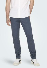 Pantalones oscuros azul marino de corte slim para hombre con una tela texturada, combinados con zapatillas blancas. Cuentan con un cierre de botón y un ligero ajuste en los tobillos.